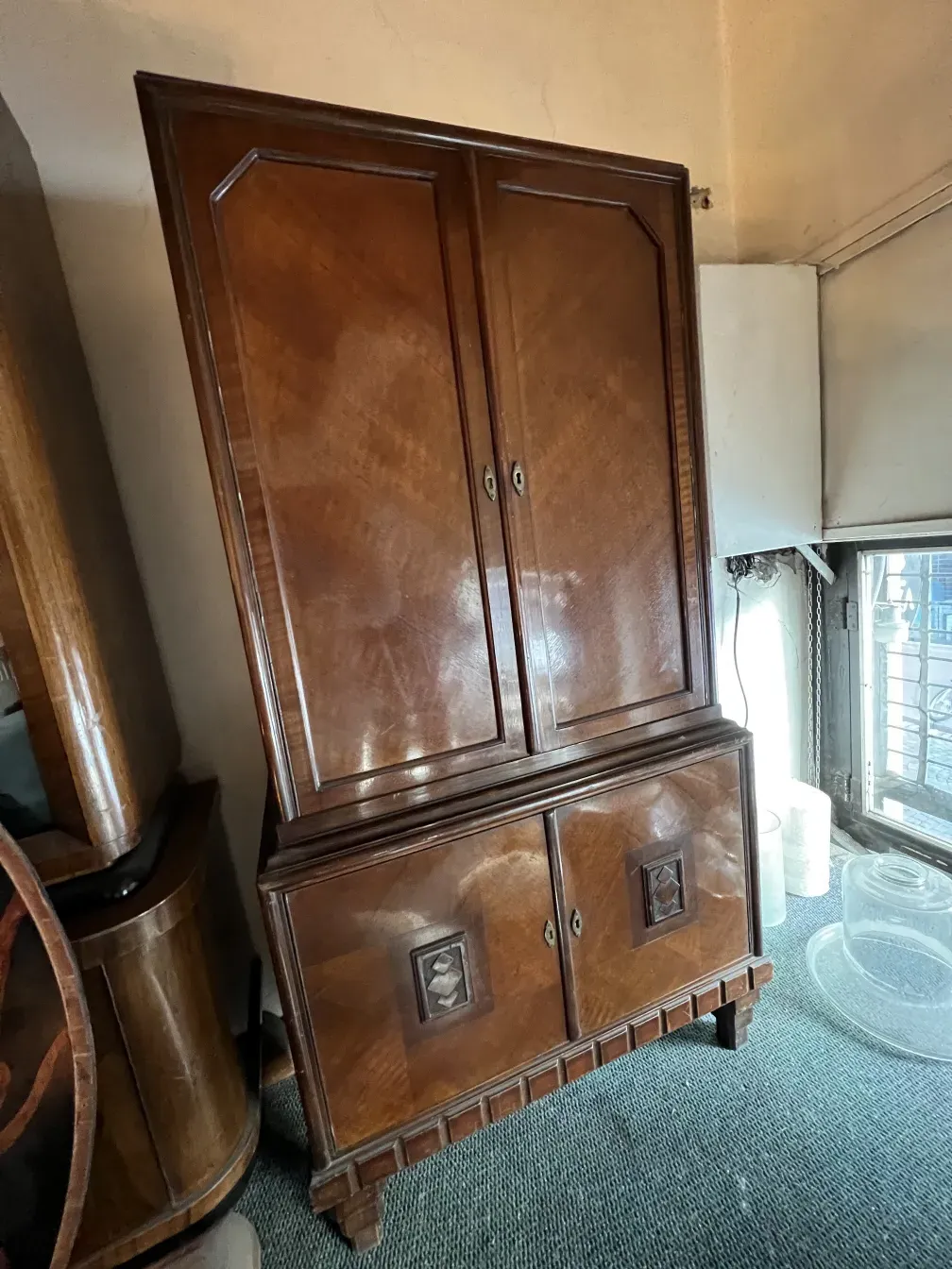 Antique wooden bookcase with intricate design and double doors, surrounded by other wooden furniture in a cozy setting.