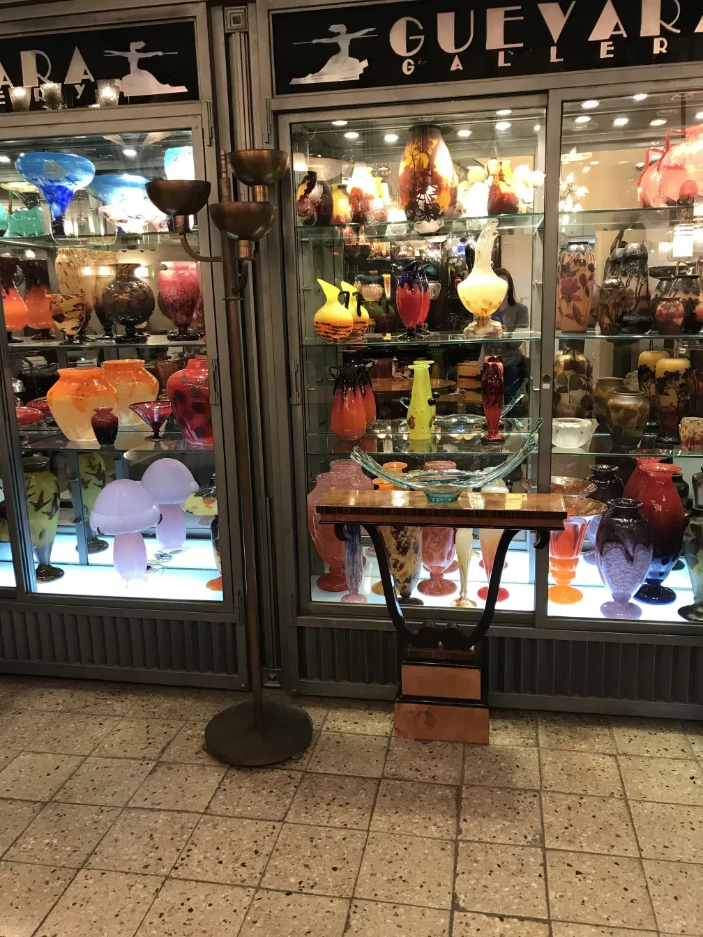 Старый торшер с классическим дизайном и двумя лампами на фоне галереи, заполненной стеклянными предметами.