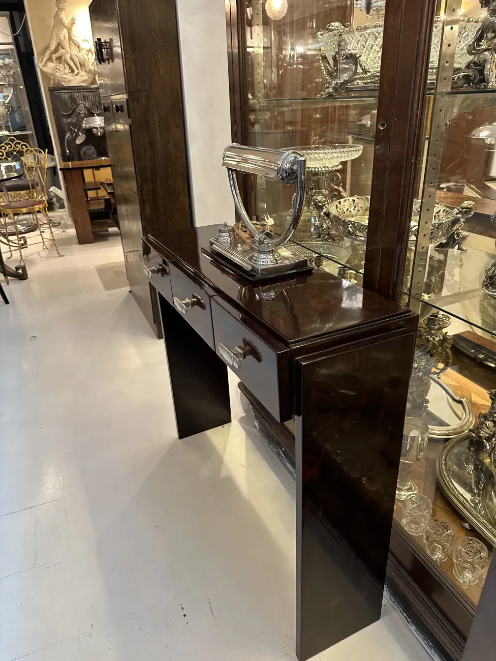 Luminária de mesa antiga com base polida e detalhes brilhantes, rodeada por objetos decorativos.