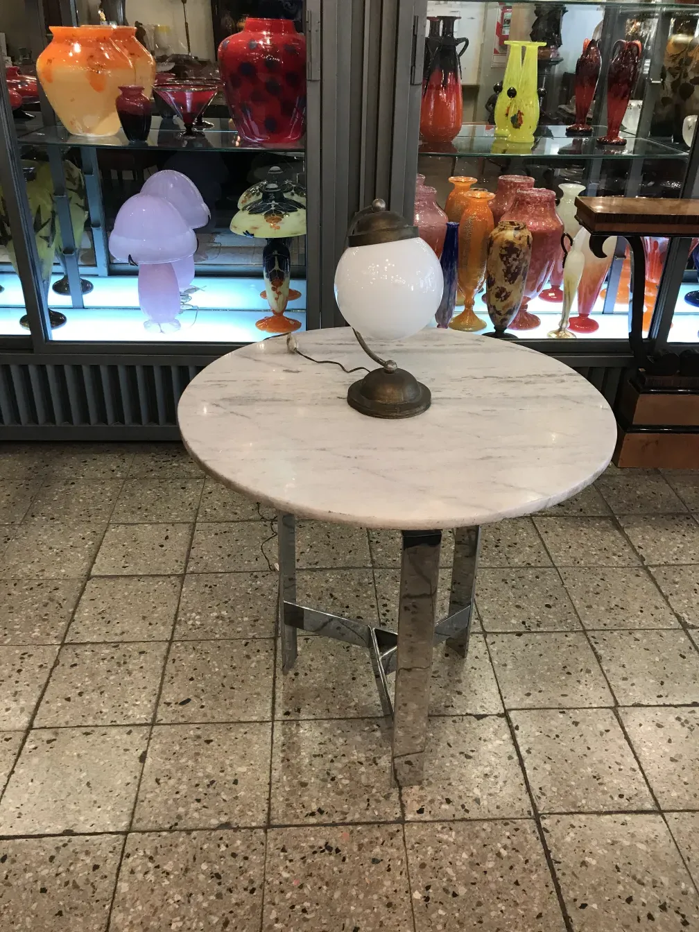 Luminária de mesa antiga com base metálica e globo branco, em meio a vasos coloridos ao fundo.