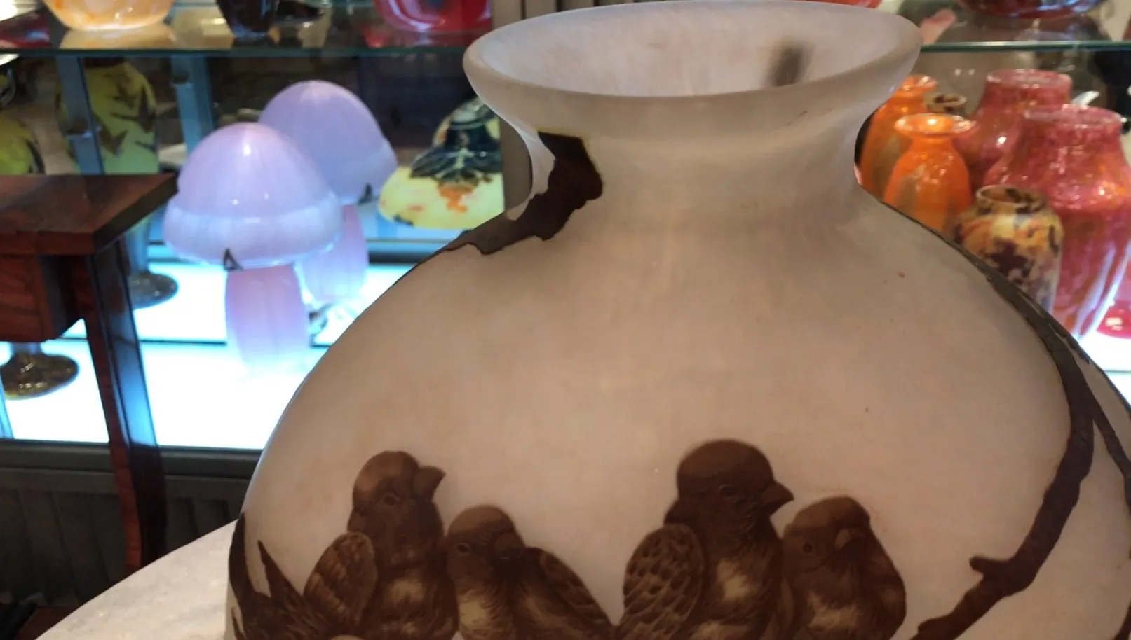 Vista de um antigo vidro assinado Muller Fres com aves decorativas, fundo inclui outros objetos de antiguidades.