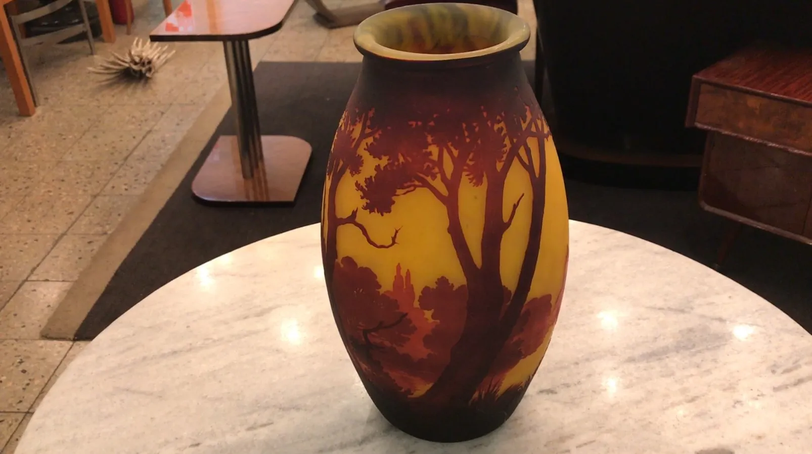 Vaso de vidro com assinatura, fundo de árvores e cores quentes.