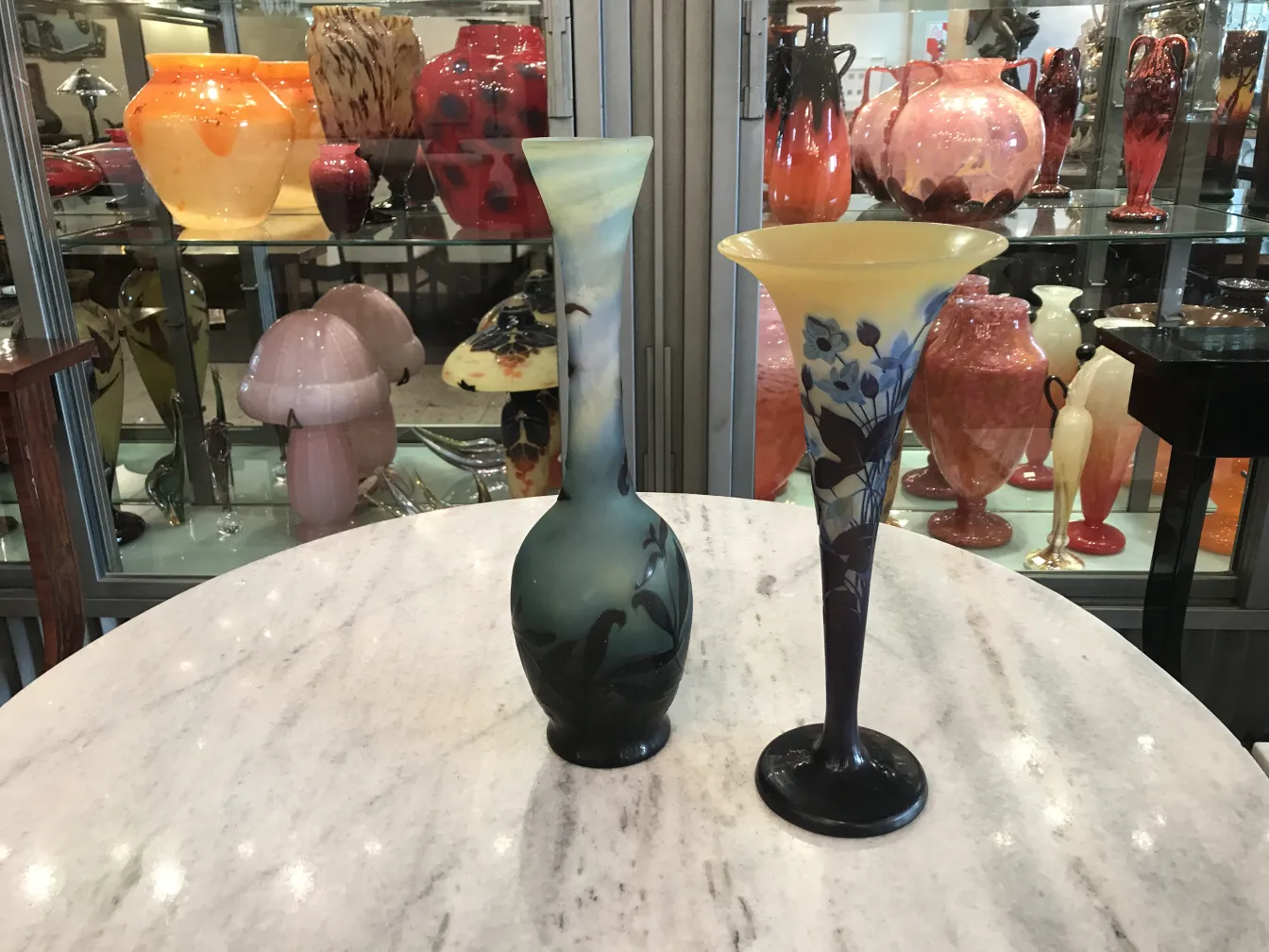 Two antique glass vases with floral decoration, one in dark tones and the other in light colors, surrounded by other antique objects.