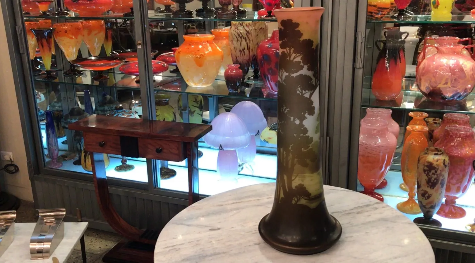 Antique vase signed Gallé with scenic design in front of a display case filled with decorative glass items.