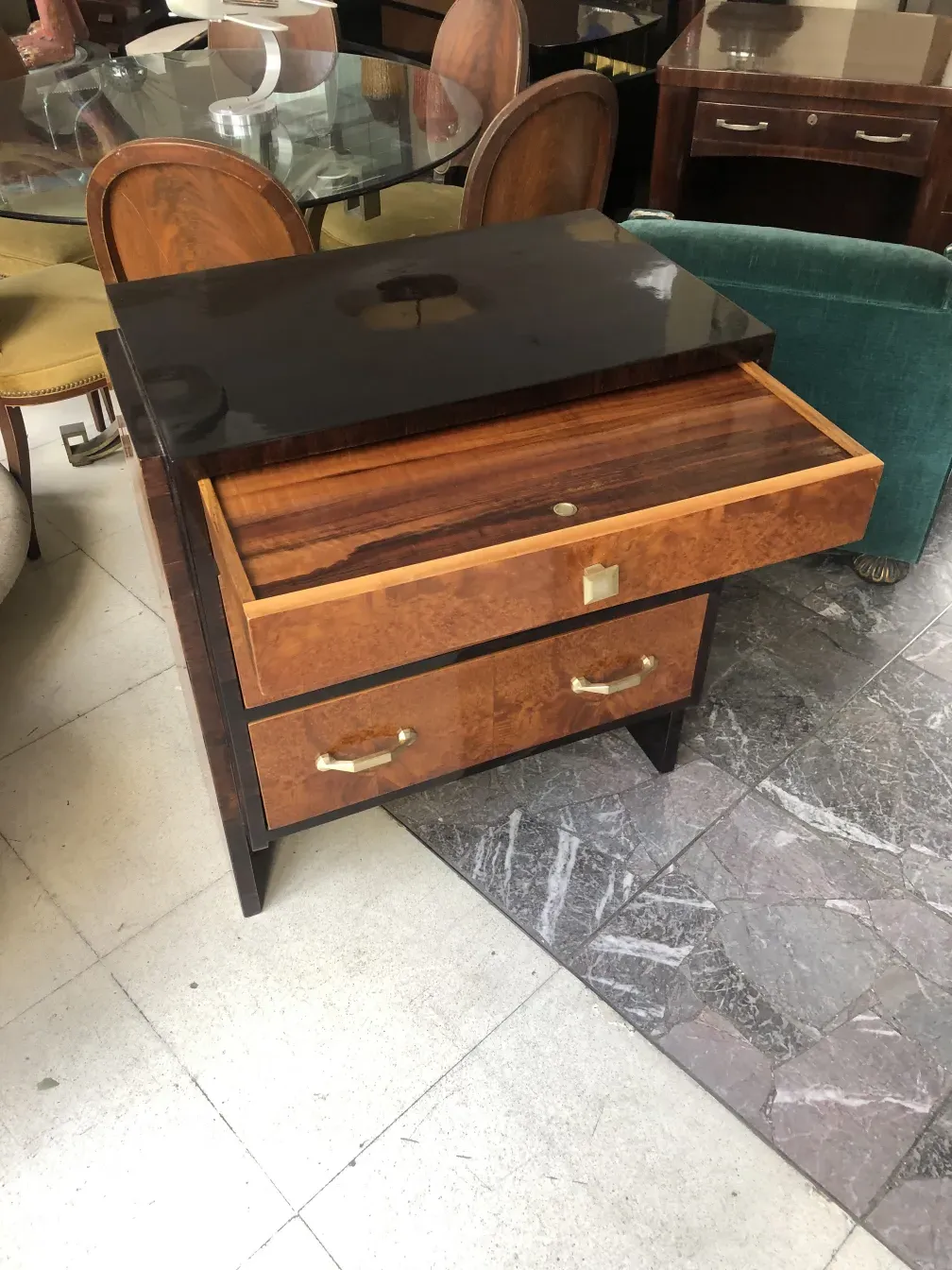 Comoda antigua de madera con dos cajones y superficie brillante, rodeada de sillas y mesas.