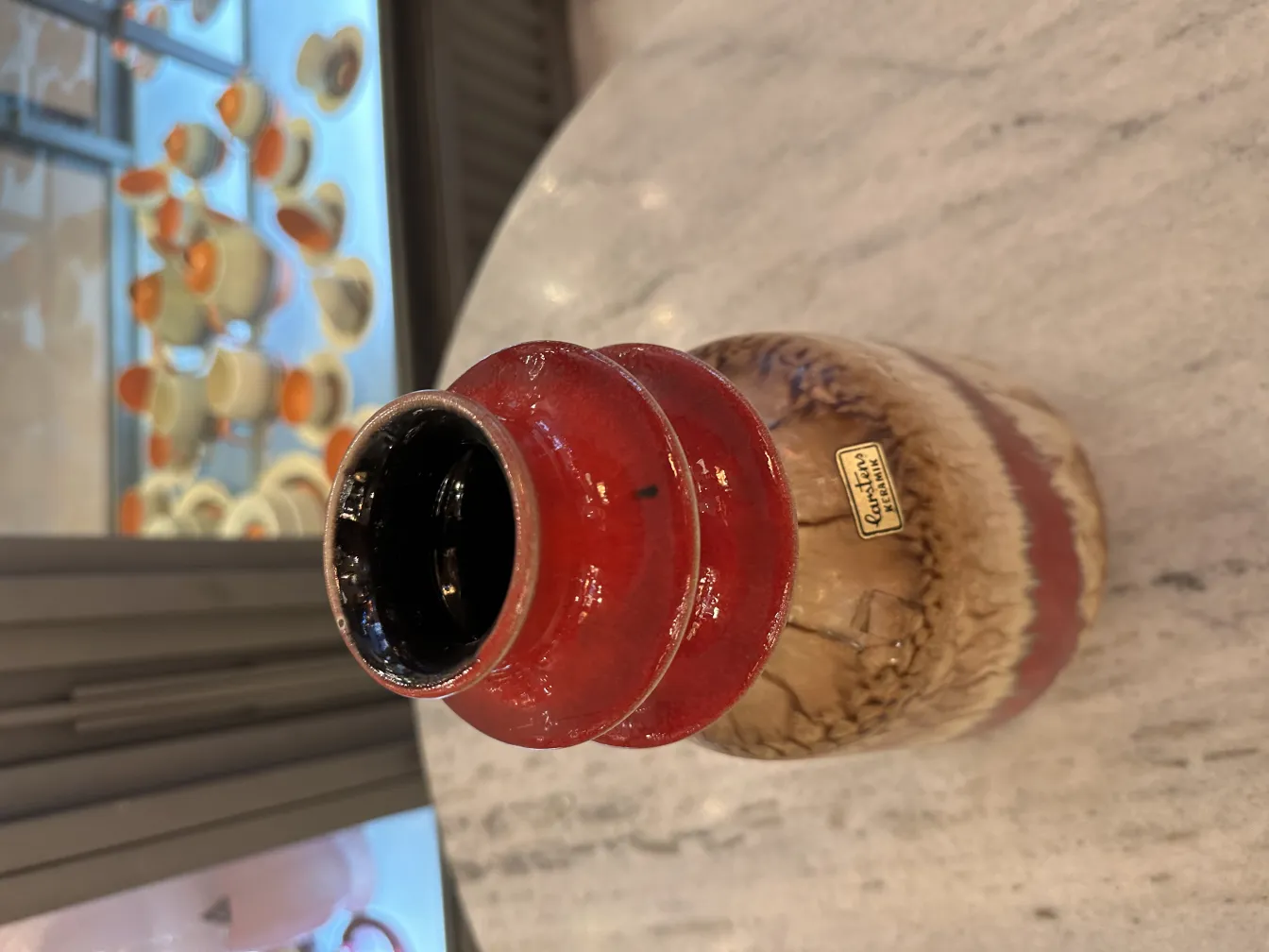 Vaso decorativo antigo de cerâmica em tons avermelhados e marrons, com fundo de xícaras brancas e laranja.