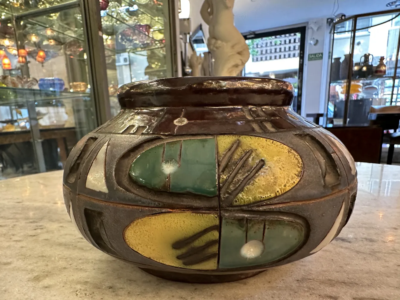 Jarrón de cerámica antigua con patrones en colores azul, verde y amarillo, sobre una mesa de mármol. Fondo con vitrinas de antigüedades.