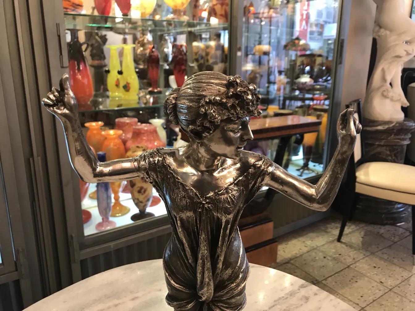 Escultura antigua de figura femenina con brazos alzados, en un ambiente lleno de jarrones coloridos.