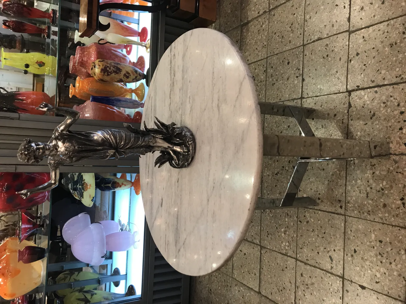 Scultura antica di figura femminile su un tavolo rotondo, con vasi colorati sullo sfondo.