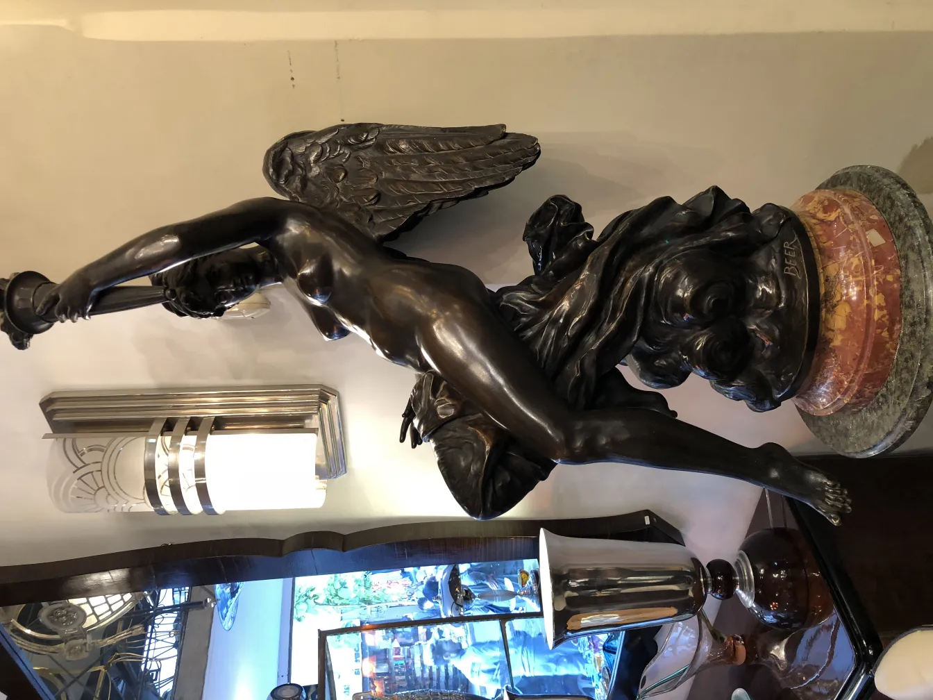 alte Skulptur einer Frau mit Flügeln und einer Laterne, dekorativer Basis und einer Bierflasche im Hintergrund