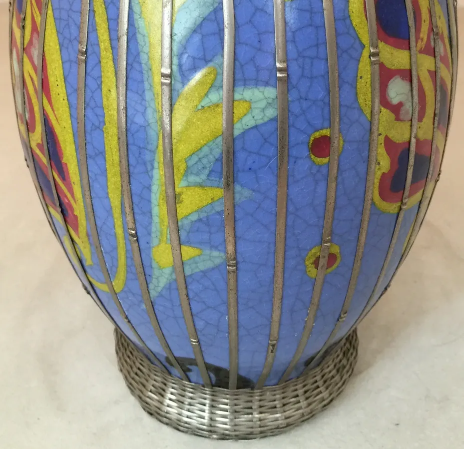 Jarrón de cerámica antigua decorada en colores vivos, con un diseño distintivo y una base de malla metálica.