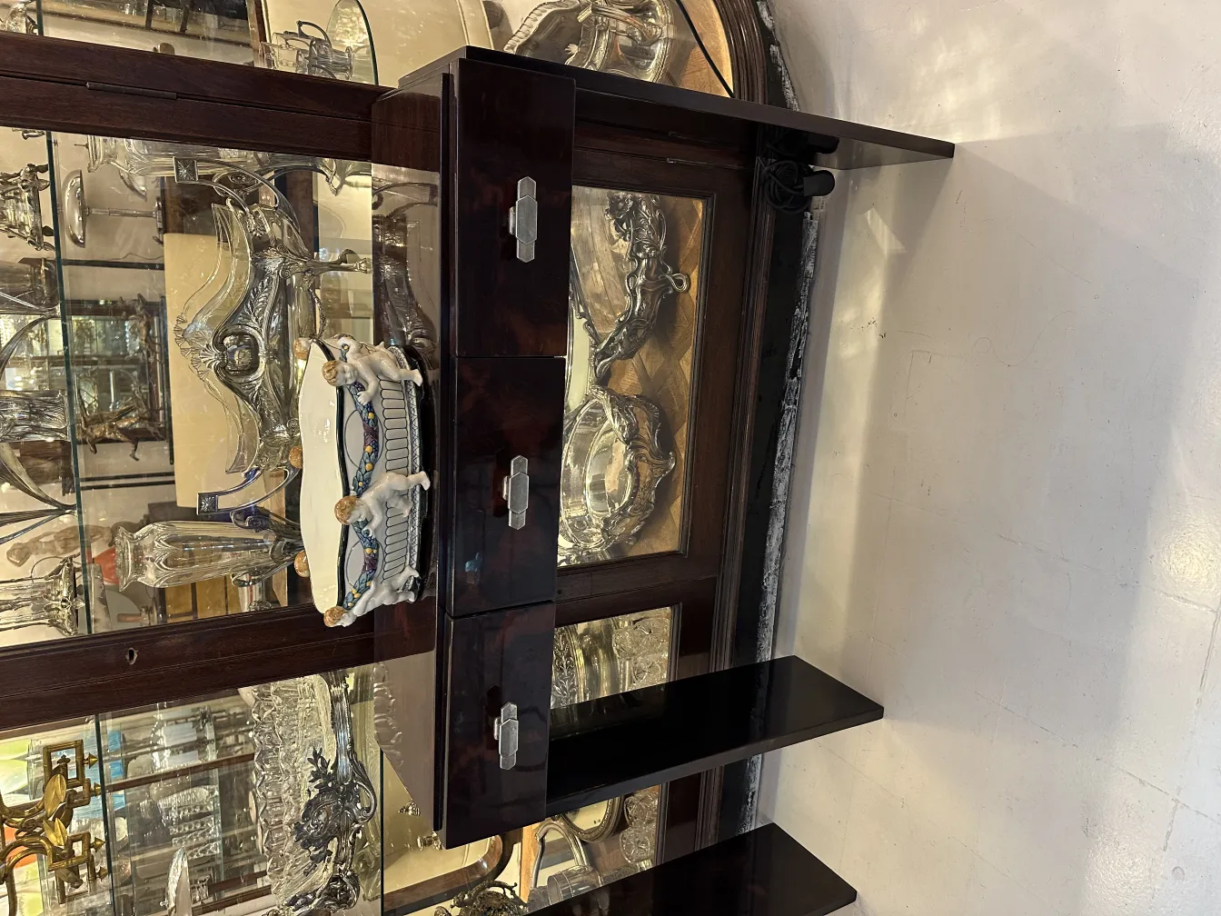 Antiga cerâmica decorativa branca com detalhes coloridos, ao fundo de objetos prateados.