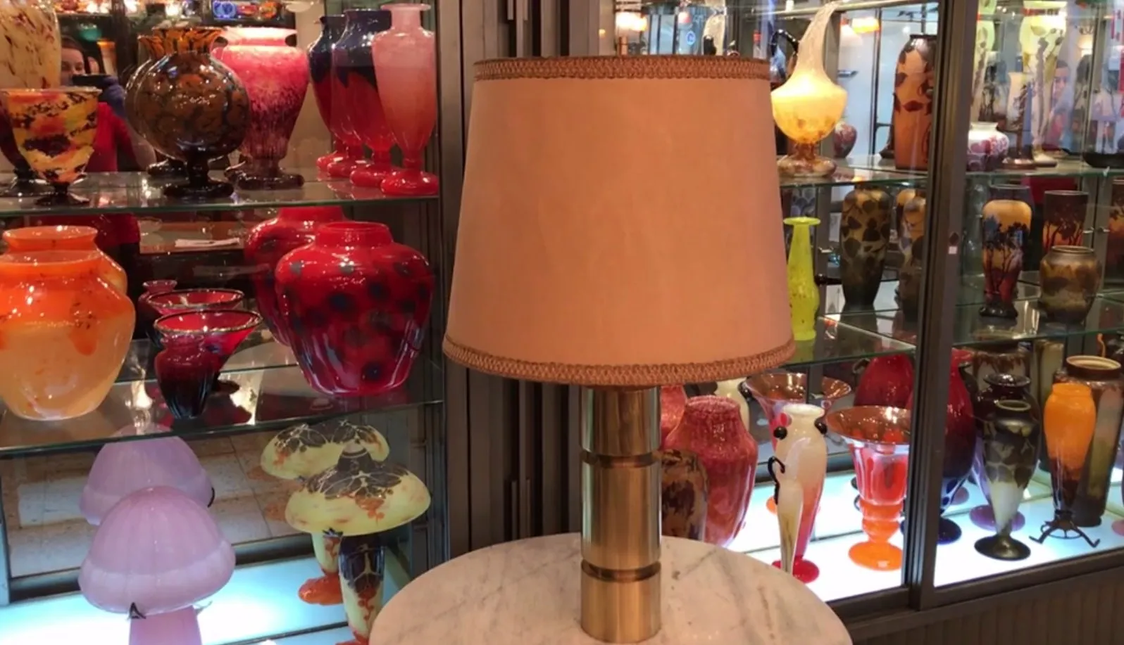 Lampe de bureau antique avec abat-jour beige dans une boutique d'antiquités entourée de vases colorés.