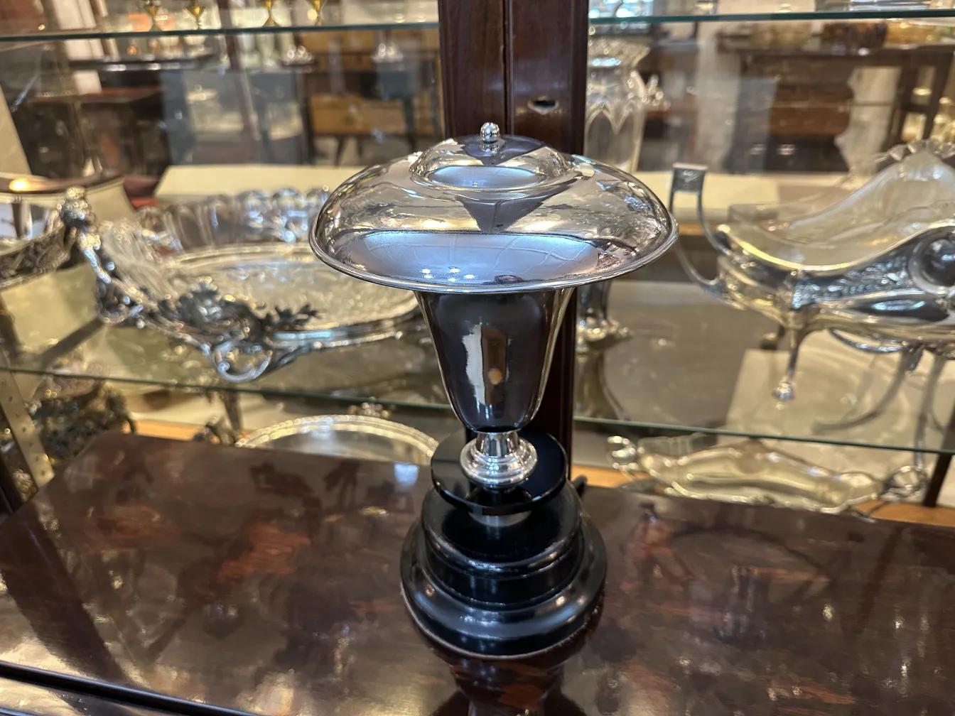 Lámpara de mesa antigua con base negra brillante y tapa plateada en un entorno de vitrina decorativa.