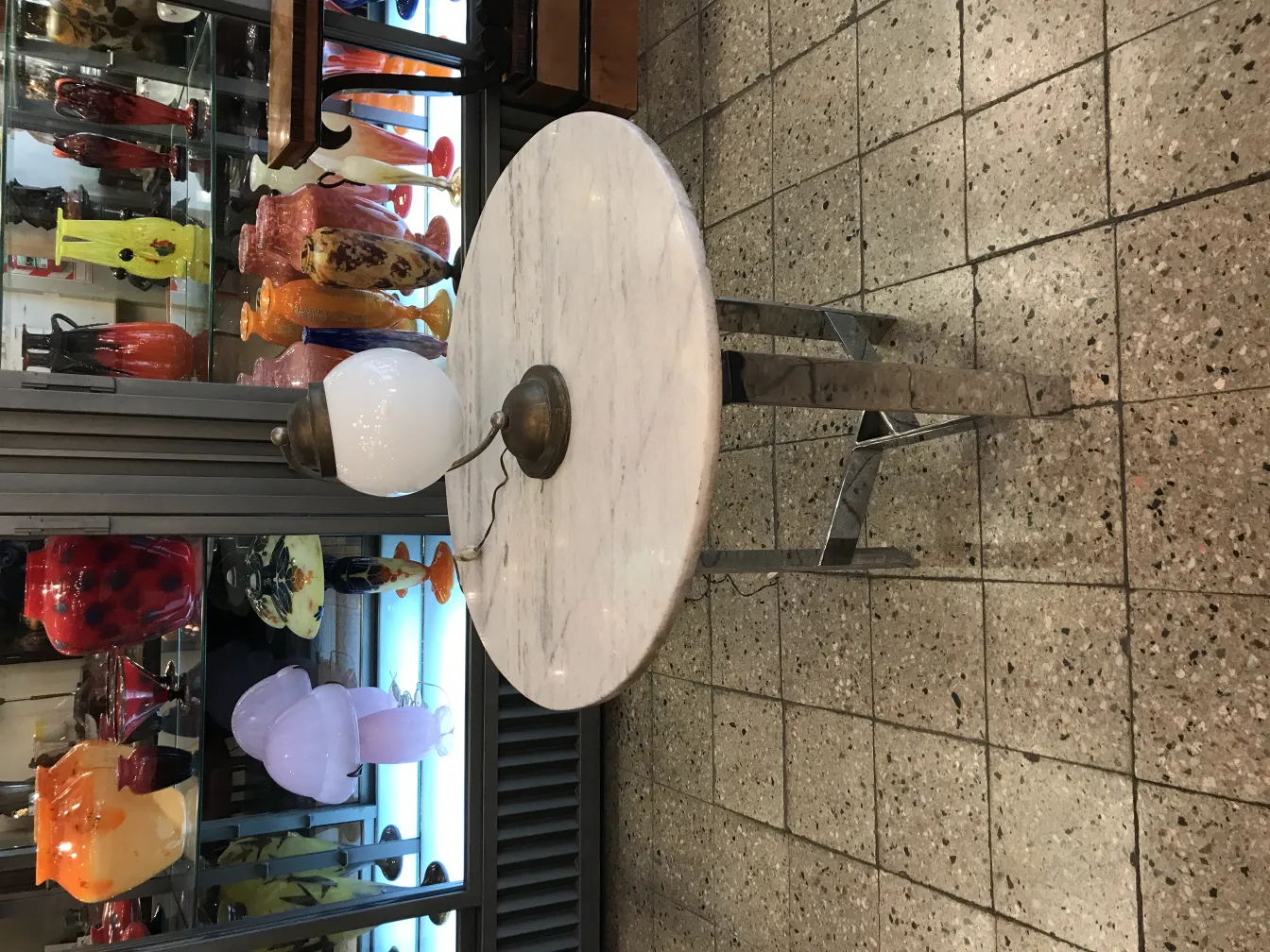 Luminária de mesa antiga com base metálica e globo branco, em meio a vasos coloridos ao fundo.