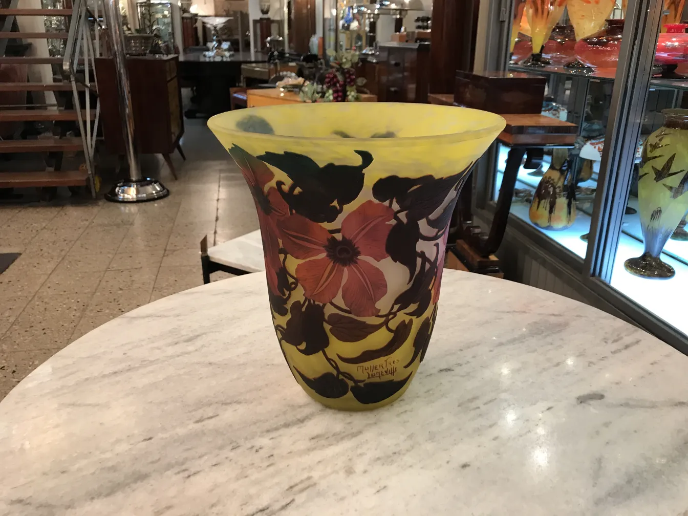 Vaso antiguo de vidrio firmado Muller Fres con flores y diseño colorido en un ambiente de antigüedades.