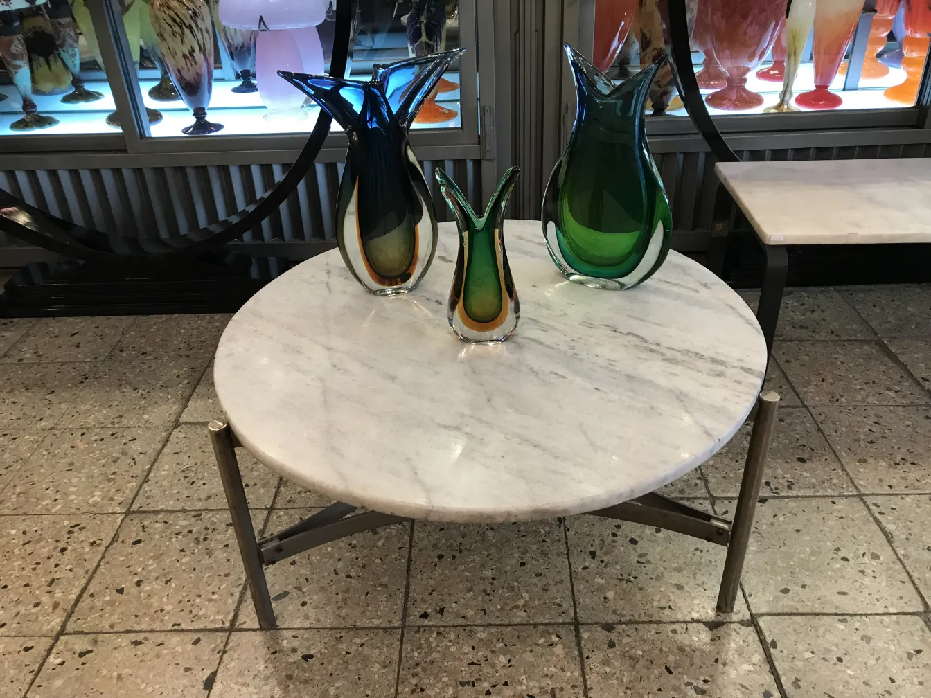 Vaso de murano em cores vibrantes sobre mesa de mármore, cercado por outros itens decorativos.