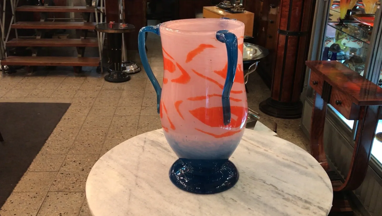 Verre décoratif signé, avec un design coloré en tons de rose et bleu, avec des poignées, sur une table en marbre.
