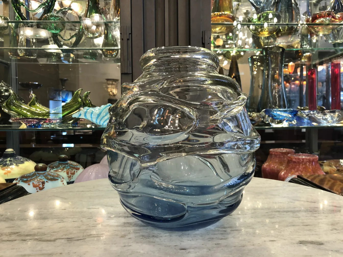 signed glass bottle in shades of blue and clear, with wavy decoration, background of antiques