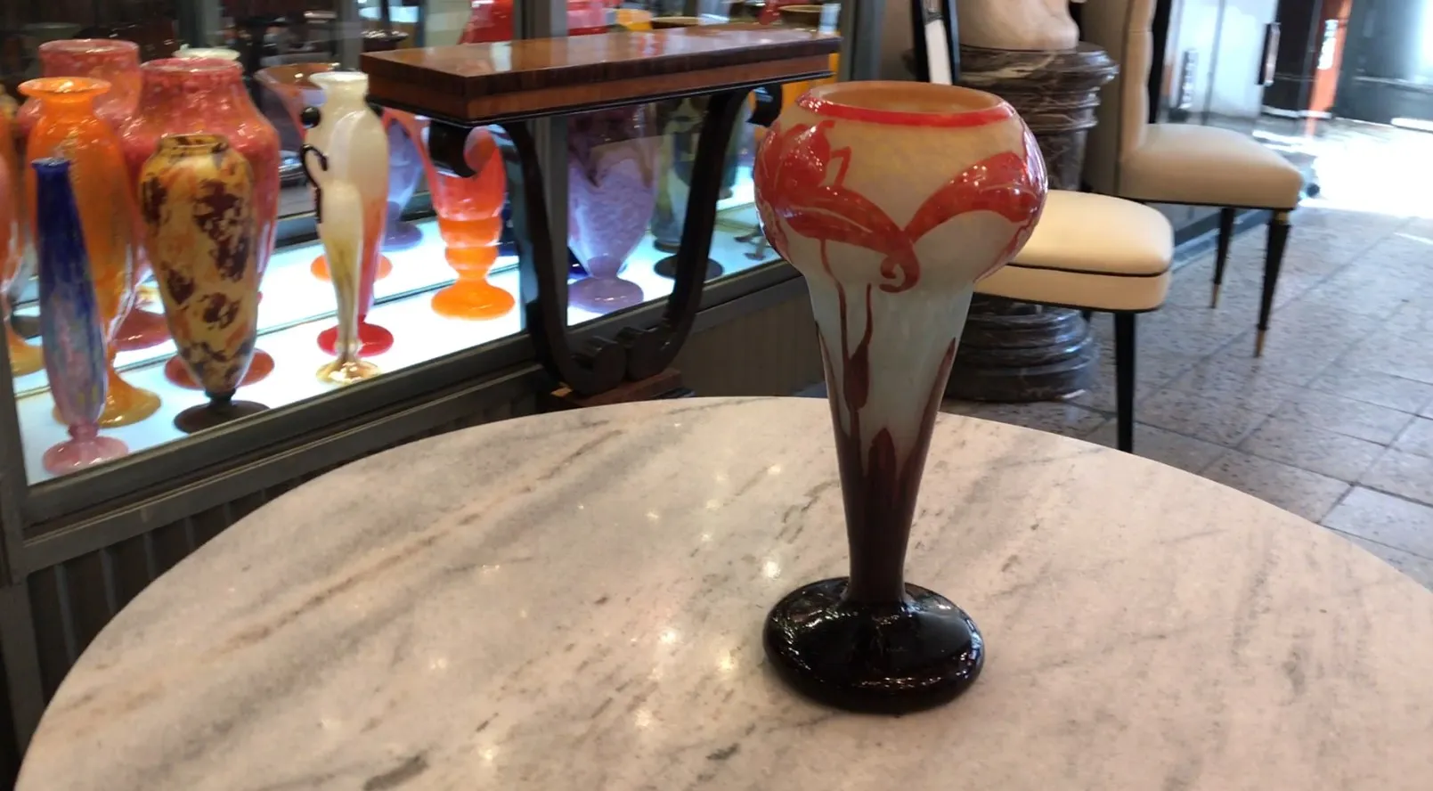 Jarrón de vidrio antiguo con diseño llamativo en rojo y blanco, fondo lleno de otros jarrones de colores.