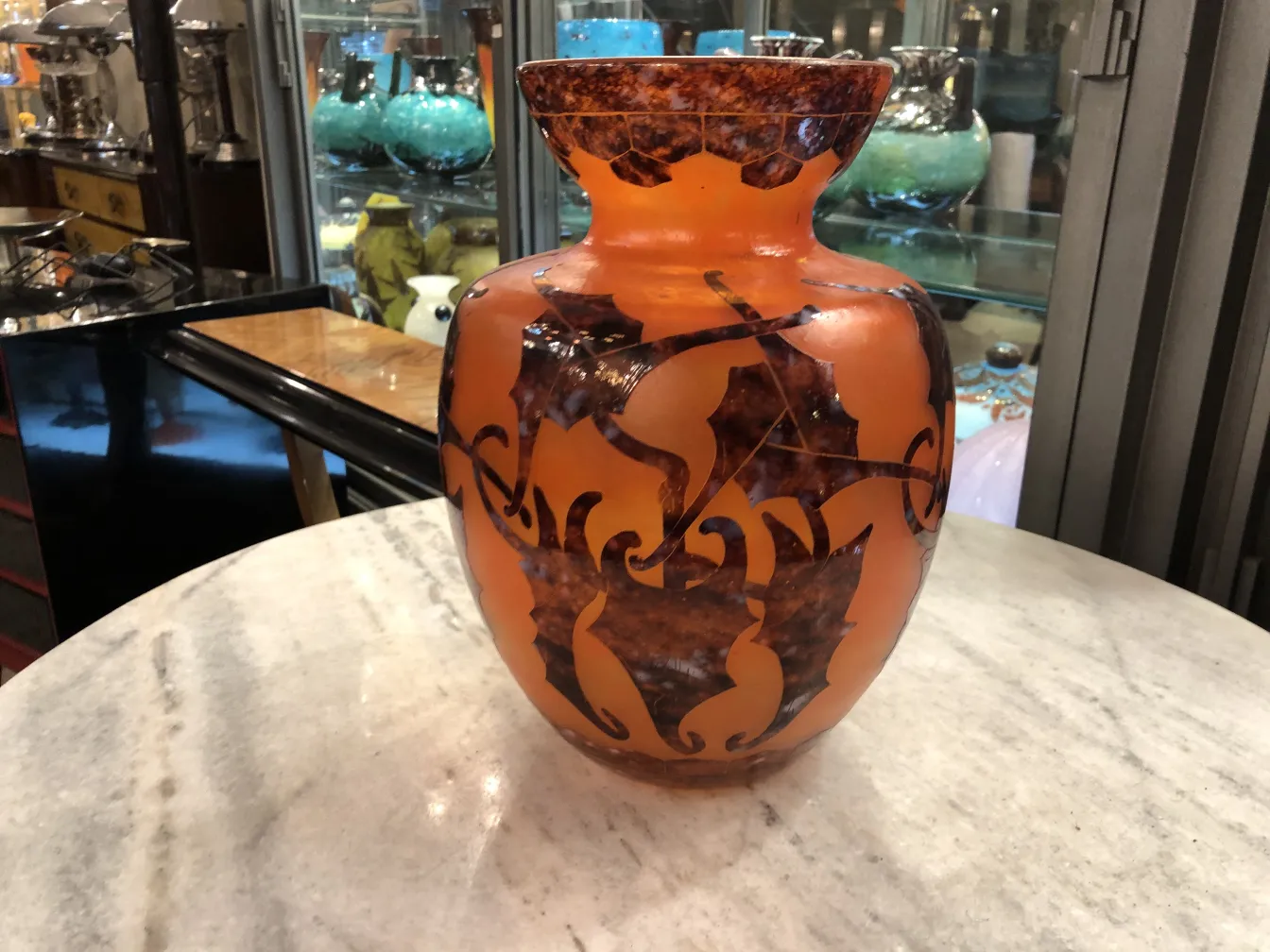 Vase en verre signé avec des détails décoratifs orange et marron, placé parmi des vitrines anciennes.