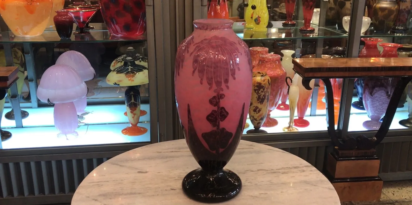 Antigo vaso de vidro com design floral em rosa, ambientado em uma vitrine cheia de outros objetos de vidro.