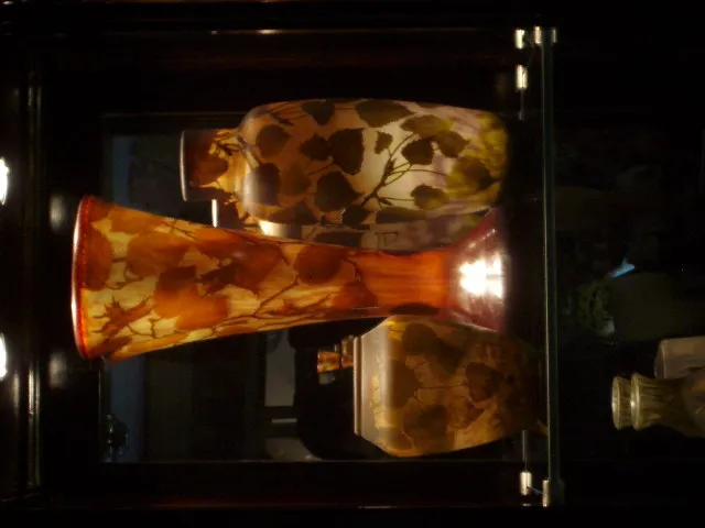 Three antique signed glass vases by Daum Nancy in warm colors, displayed in a showcase. Elegant setting.