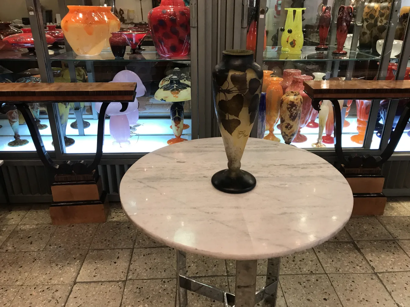 Vaso decorativo de vidro assinado Daum Nancy com folhas, iluminado por trás com outros itens de vidro colorido.