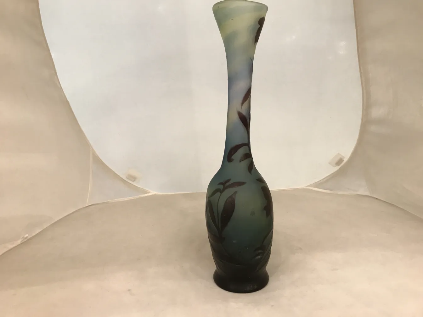 Un ancien vase en verre avec la signature de Gallé, orné de motifs floraux dans des tons verts et bleus sur un fond clair.