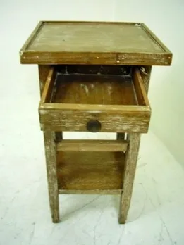 Antigua mesita con un cajón, con un diseño sencillo y funcional, situada en un ambiente neutro.