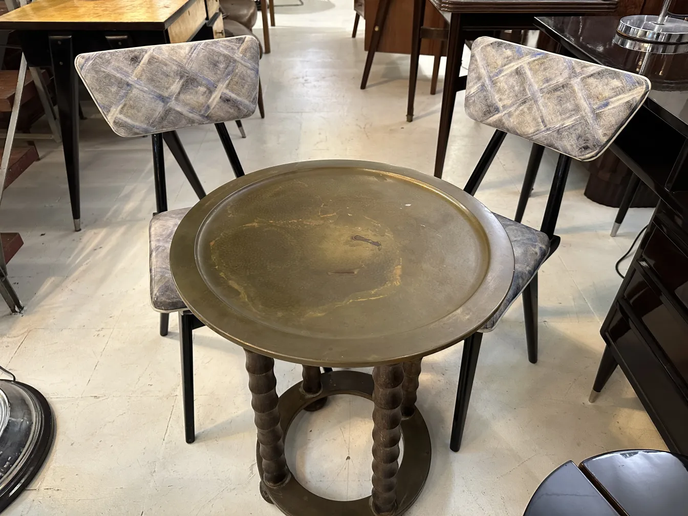 Petite table ancienne avec base tordue et plateau doré, entourée de chaises. Ambiance chaleureuse.
