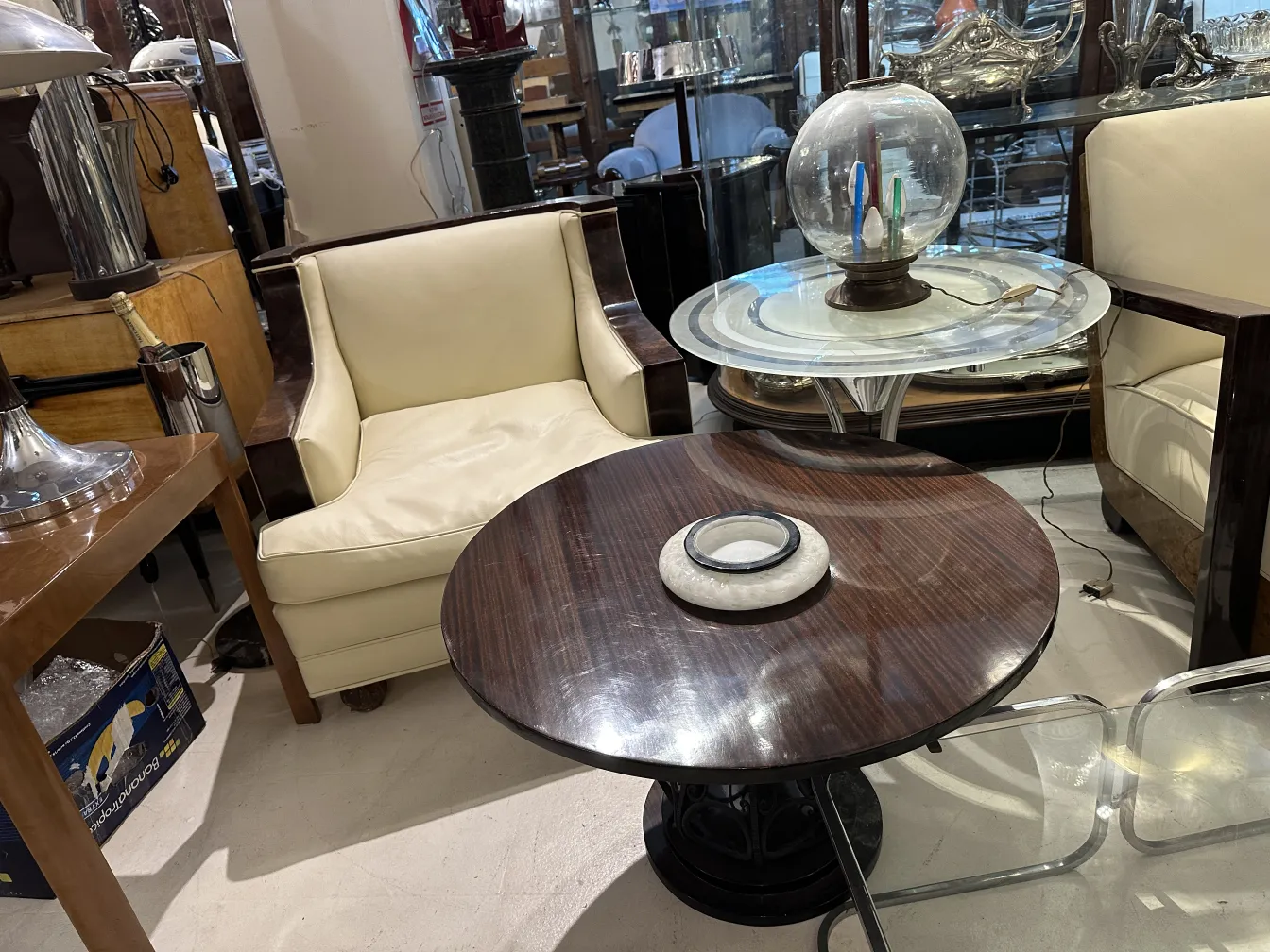 mesita antigua con un diseño redondo y base decorativa, en un entorno elegante con sillas claras.