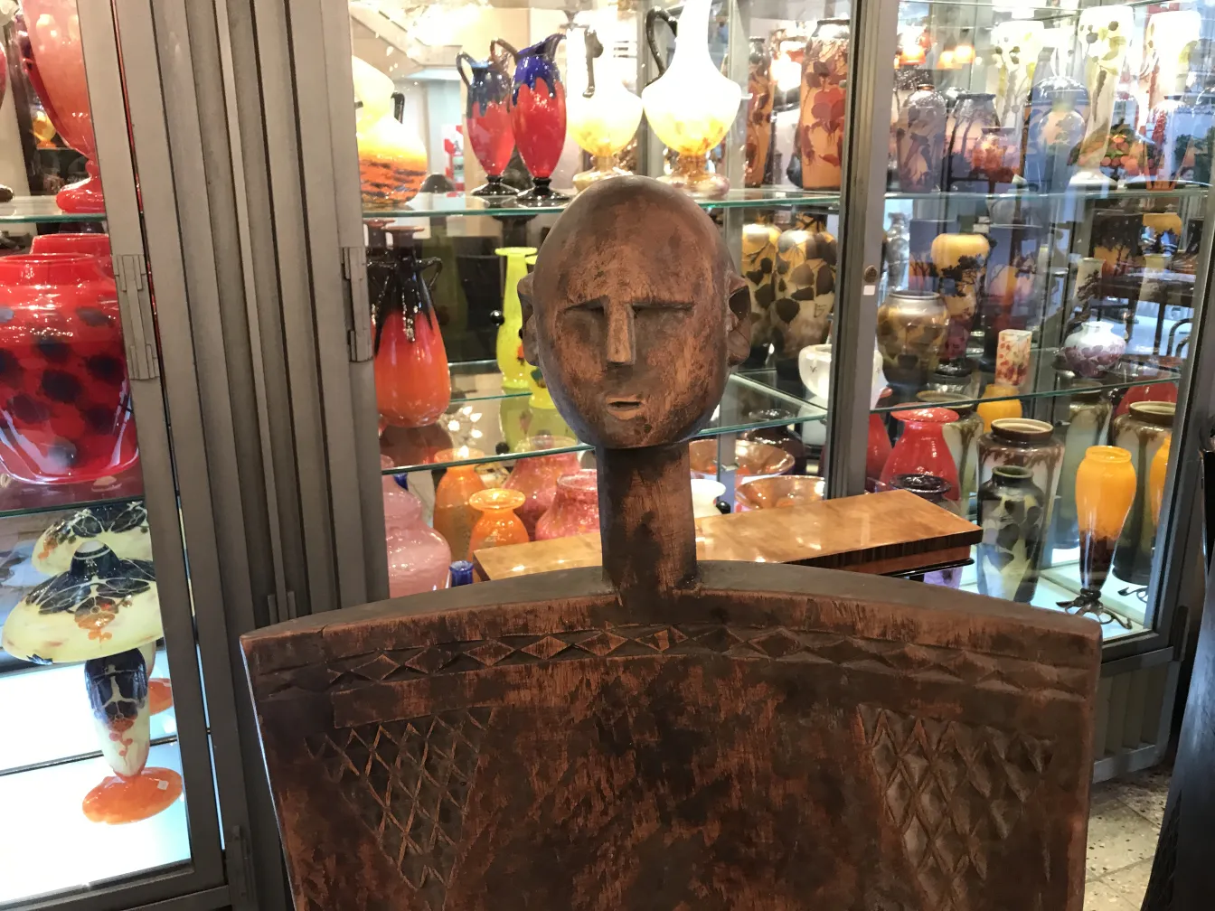 Due sedie antiche con design tribale e schienale alto, con figura maschile, sullo sfondo di oggetti decorativi colorati.