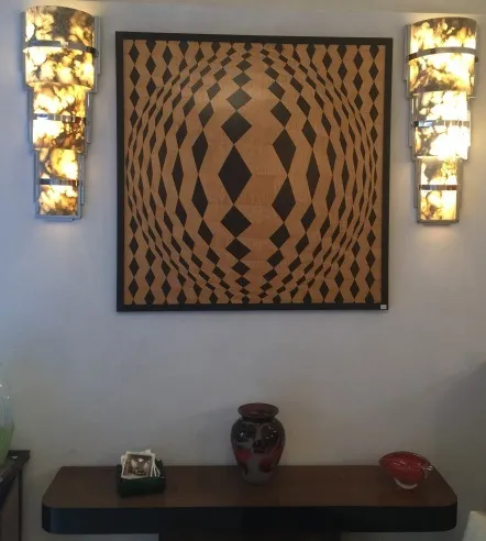 Antigua marquetería decorativa con patrones geométricos en tonos oscuros y claros, enmarcada elegantemente en la pared, junto a luces. Un jarrón y un plato decorativo en la mesa.