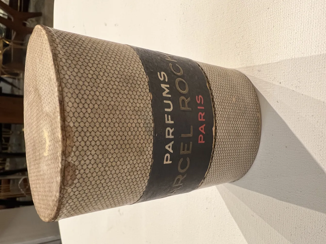 Frasco de perfume antigo de forma cilíndrica com etiqueta de Paris, fundo claro.