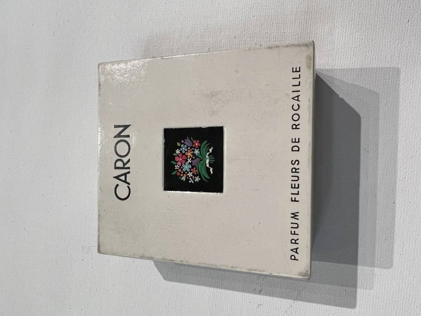 Flacon de parfum ancien avec design floral, élégant et décoratif, forme rectangulaire, fond blanc avec des détails noirs et colorés.