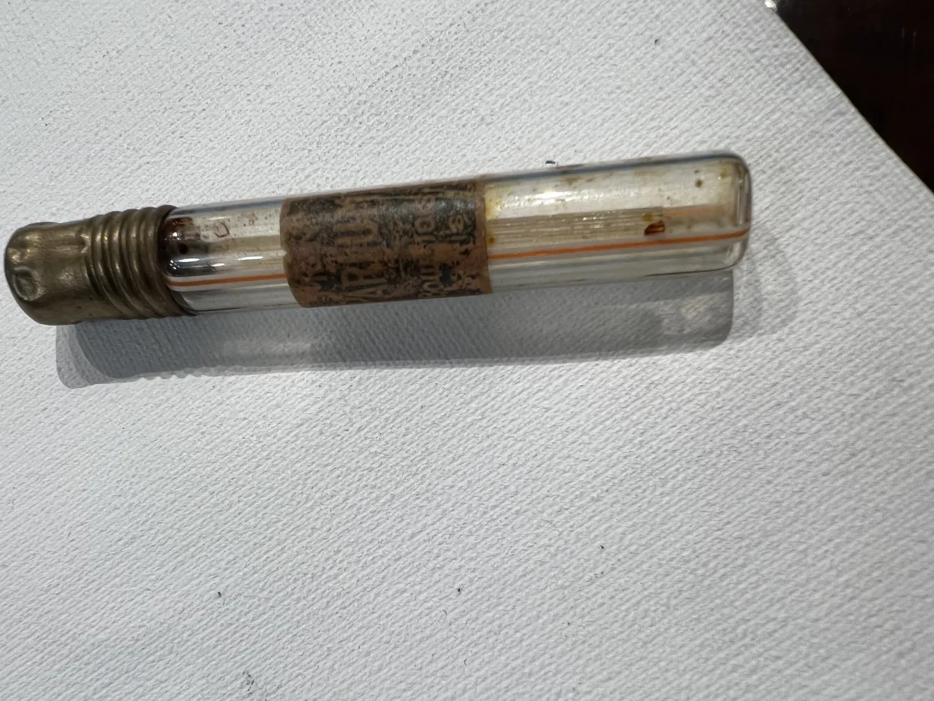 Frasco de perfume antiguo con tapa de metal, decorado con etiquetas y colores claros, sobre fondo blanco.