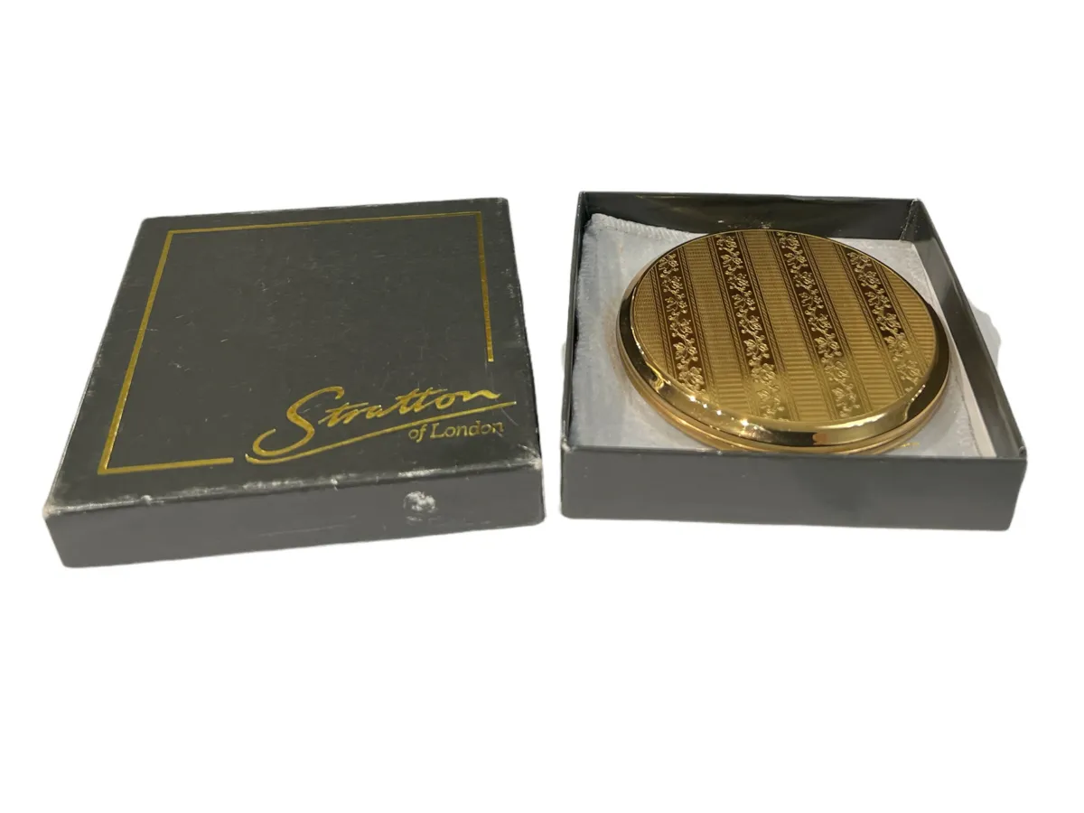 Polvera antigua con un acabado dorado y detalles decorativos, presentada en una caja negra.