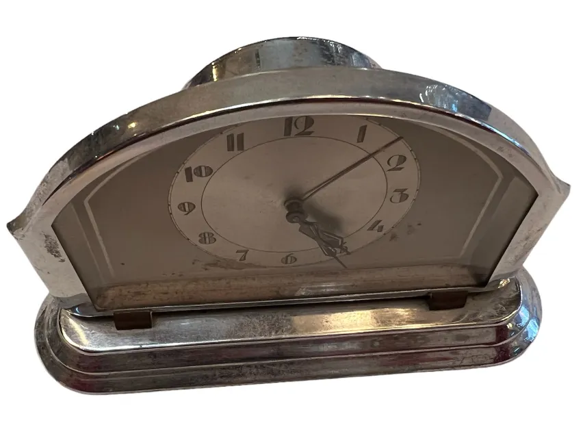 Horloge de table ancienne avec un design arqué et des aiguilles visibles, placée sur une surface claire. Fond flou avec une ambiance calme.