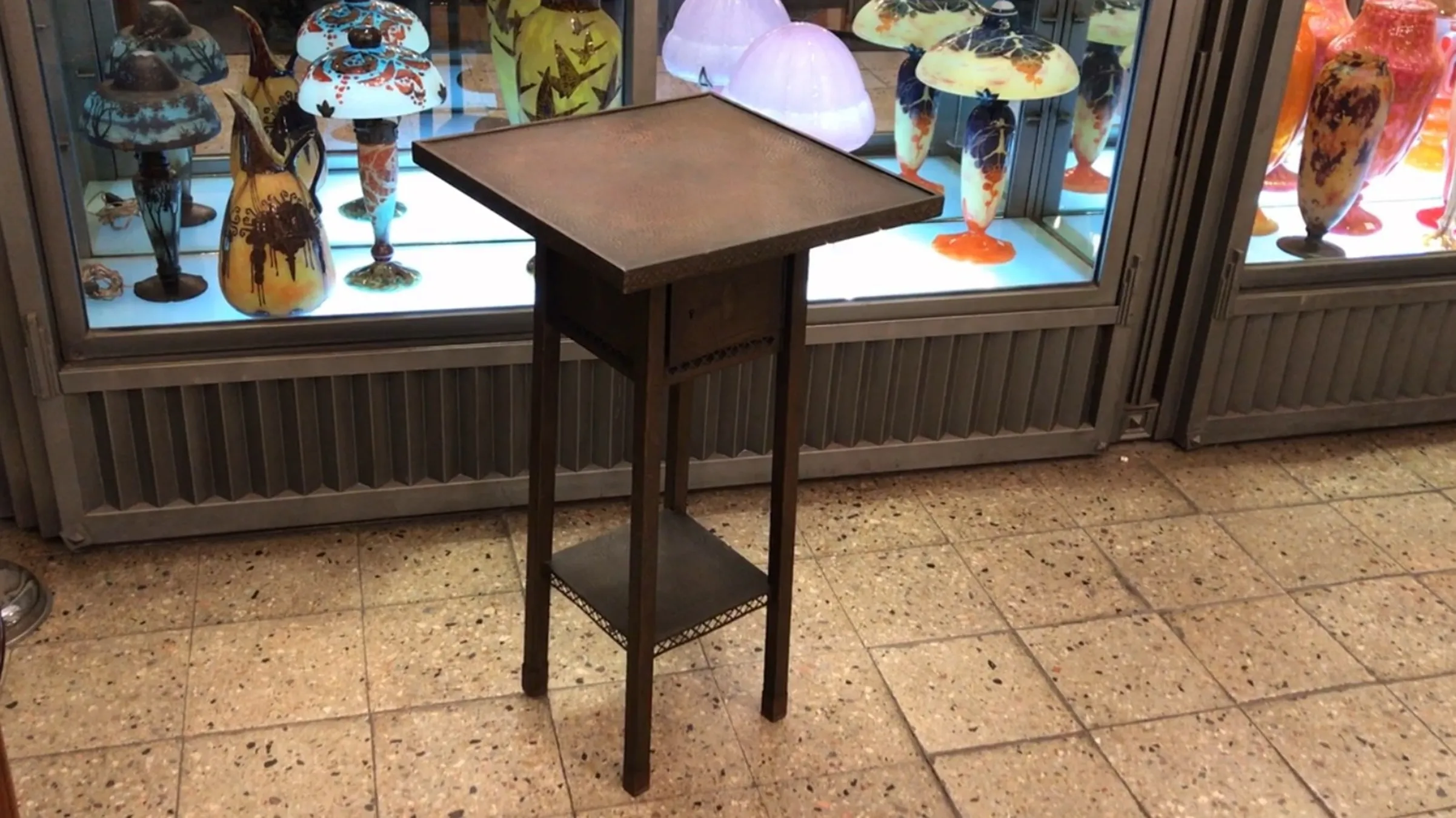 Mesita antigua pequeña con detalles decorativos, en un entorno de antigüedades.