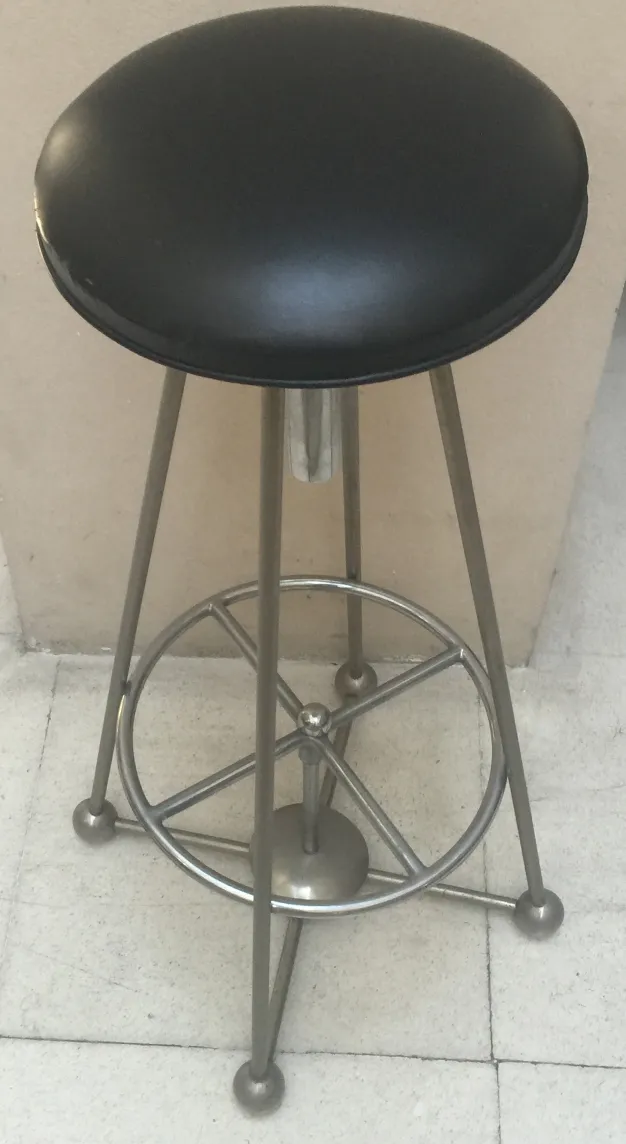 Antiga banqueta com assento redondo preto e base prateada contra um fundo simples.