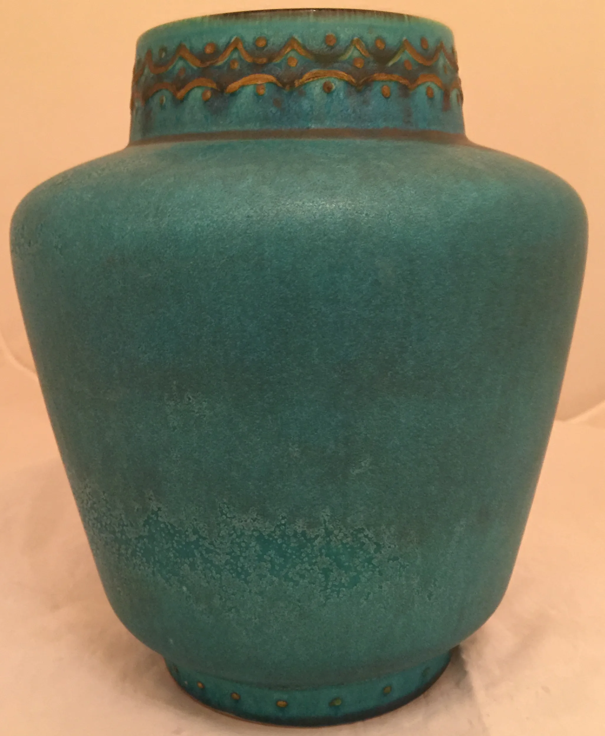 Vase en céramique antique de couleur turquoise, avec des détails décoratifs en haut, sur fond clair.