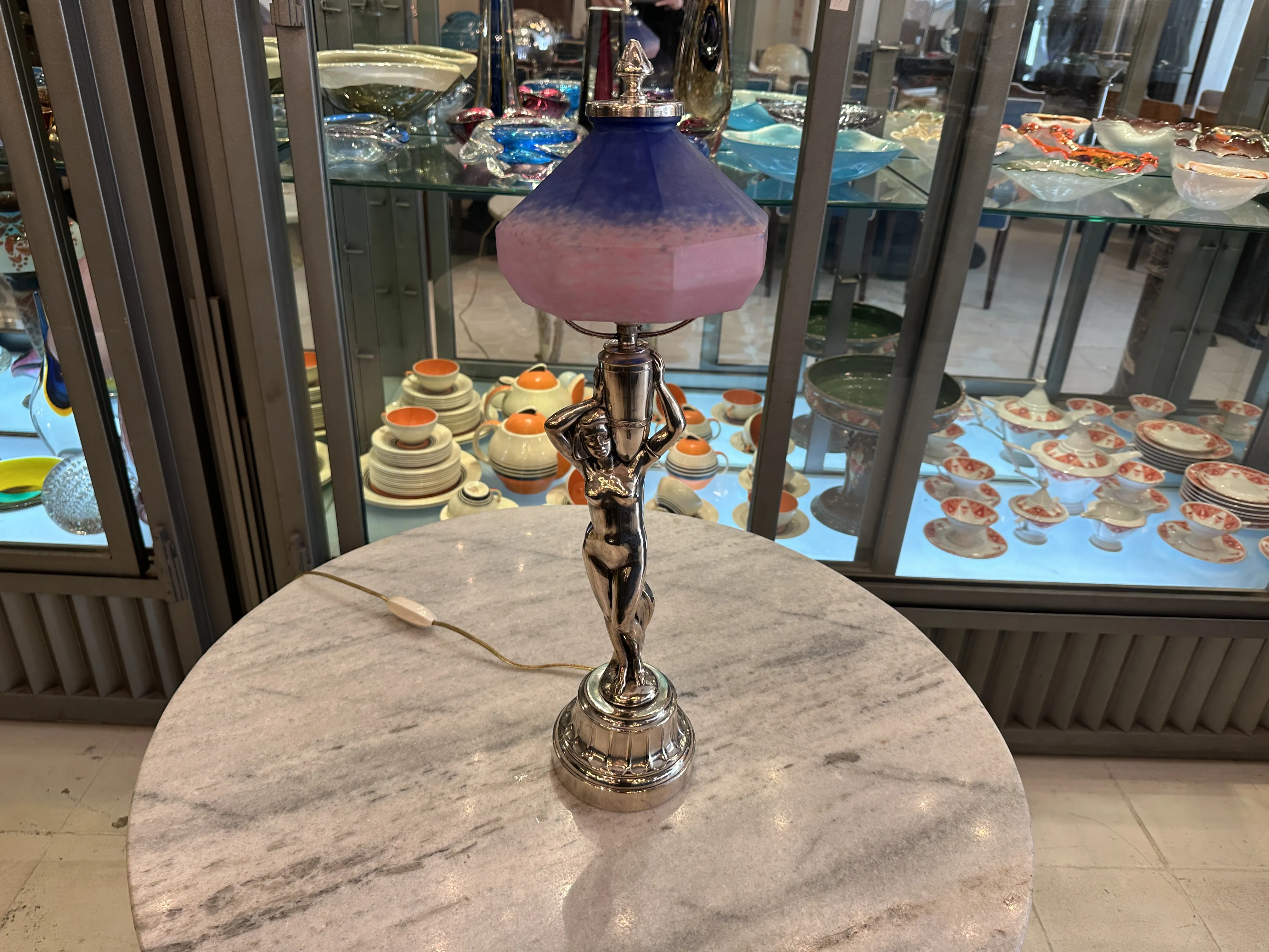 Antiga luminária de mesa com base brilhante e abajur colorido, ambientada em um espaço elegante com objetos decorativos.